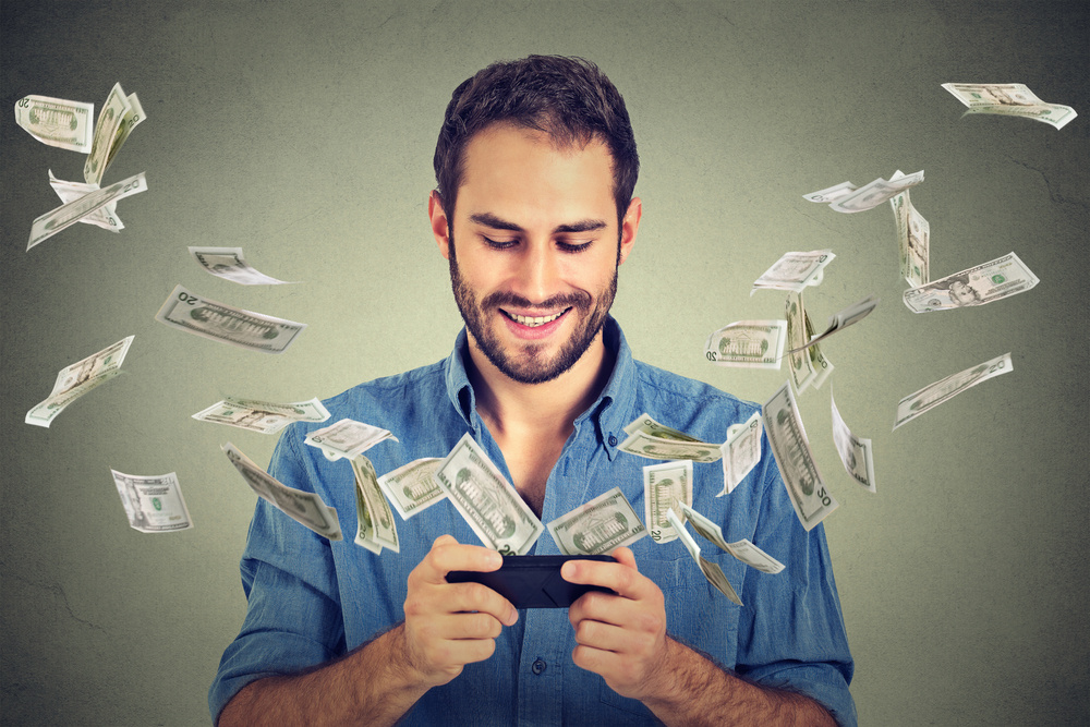 Technology online banking money transfer, e-commerce concept. Happy young man using smartphone with dollar bills flying away from screen isolated on gray wall office background. Technology online banking money transfer, e-commerce concept. Happy young man using smartphone with dollar bills flying away from screen isolated on gray wall office background.